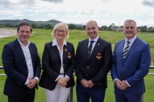 Four people in business attire posing on a golf course, smiling with greens and hills in the background.