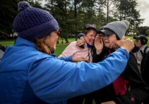 Group of people outdoors sharing a warm hug and laugh in a park, wearing jackets and hats.