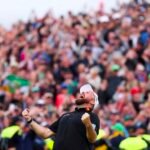 Male athlete in a black polo with a white cap celebrates with fists raised as a large cheering crowd watches.