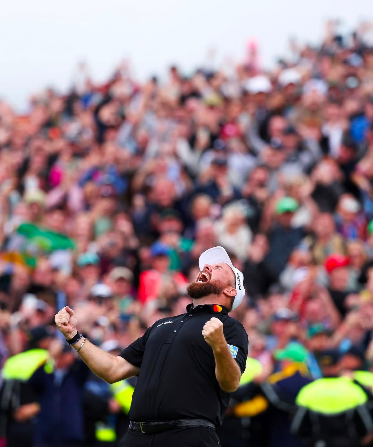 Male athlete in a black polo with a white cap celebrates with fists raised as a large cheering crowd watches.