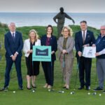 Seven professionals in business attire stand with golf clubs on a seaside putting green, holding a Kerry sign (discoverkerry.com).