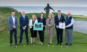Seven professionals in business attire stand with golf clubs on a seaside putting green, holding a Kerry sign (discoverkerry.com).