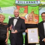 Four formally dressed people on stage at an awards ceremony; man holds a crystal trophy, woman holds a framed certificate, smiling backdrop with a Killarney crest behind them.