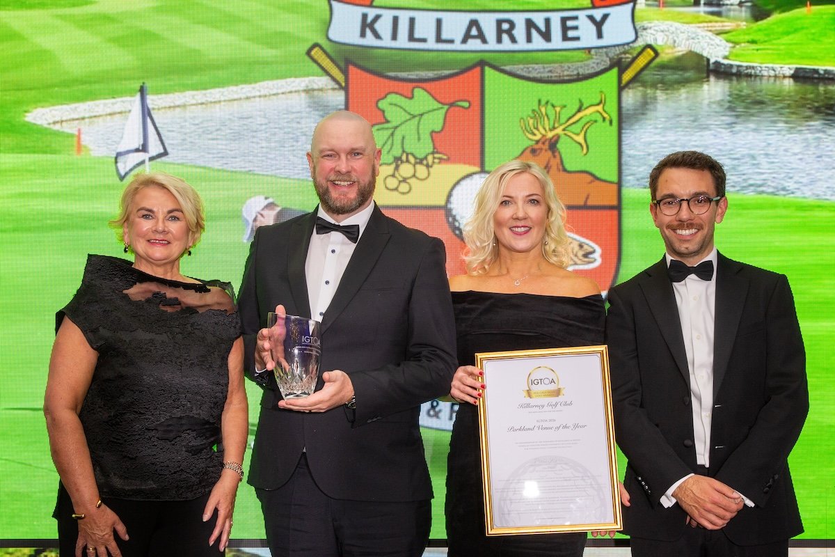 Four formally dressed people on stage at an awards ceremony; man holds a crystal trophy, woman holds a framed certificate, smiling backdrop with a Killarney crest behind them.